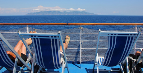 Traversée en mer d’un ferry de la compagnie maritime Moby Lines vers la Sardaigne avec trois transats bleus avec des rayures blanches verticales, un filet blanc sur les barrières arrière du bateau avec deux personnes prenant le soleil allongé sur les transats et les pieds sur la barrière blanche.